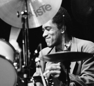 Photo noir et blanc du batteur Al Foster, souriant et en train de jouer sur scène (site de vente de vinyles jazz Everything jazz).