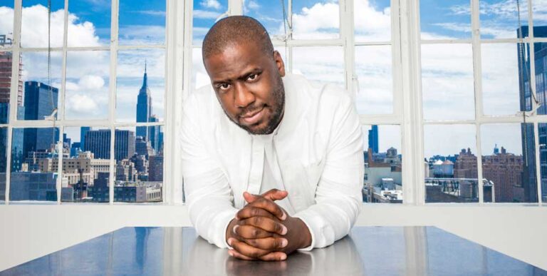 Portrait photo couleur de Robert Glasper en costume blanc devant une large fenêtre avec vue sur des gratte-ciel par temps clair et nuageux A découvrir sur EverythingJazz, site de vente de vinyles.