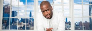 Portrait photo couleur de Robert Glasper en costume blanc devant une large fenêtre avec vue sur des gratte-ciel par temps clair et nuageux A découvrir sur EverythingJazz, site de vente de vinyles.