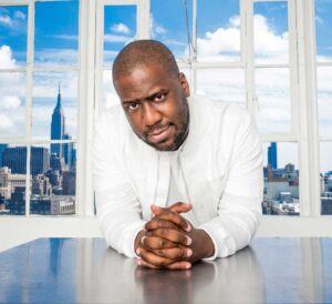 Portrait photo couleur de Robert Glasper en costume blanc devant une large fenêtre avec vue sur des gratte-ciel par temps clair et nuageux A découvrir sur EverythingJazz, site de vente de vinyles.